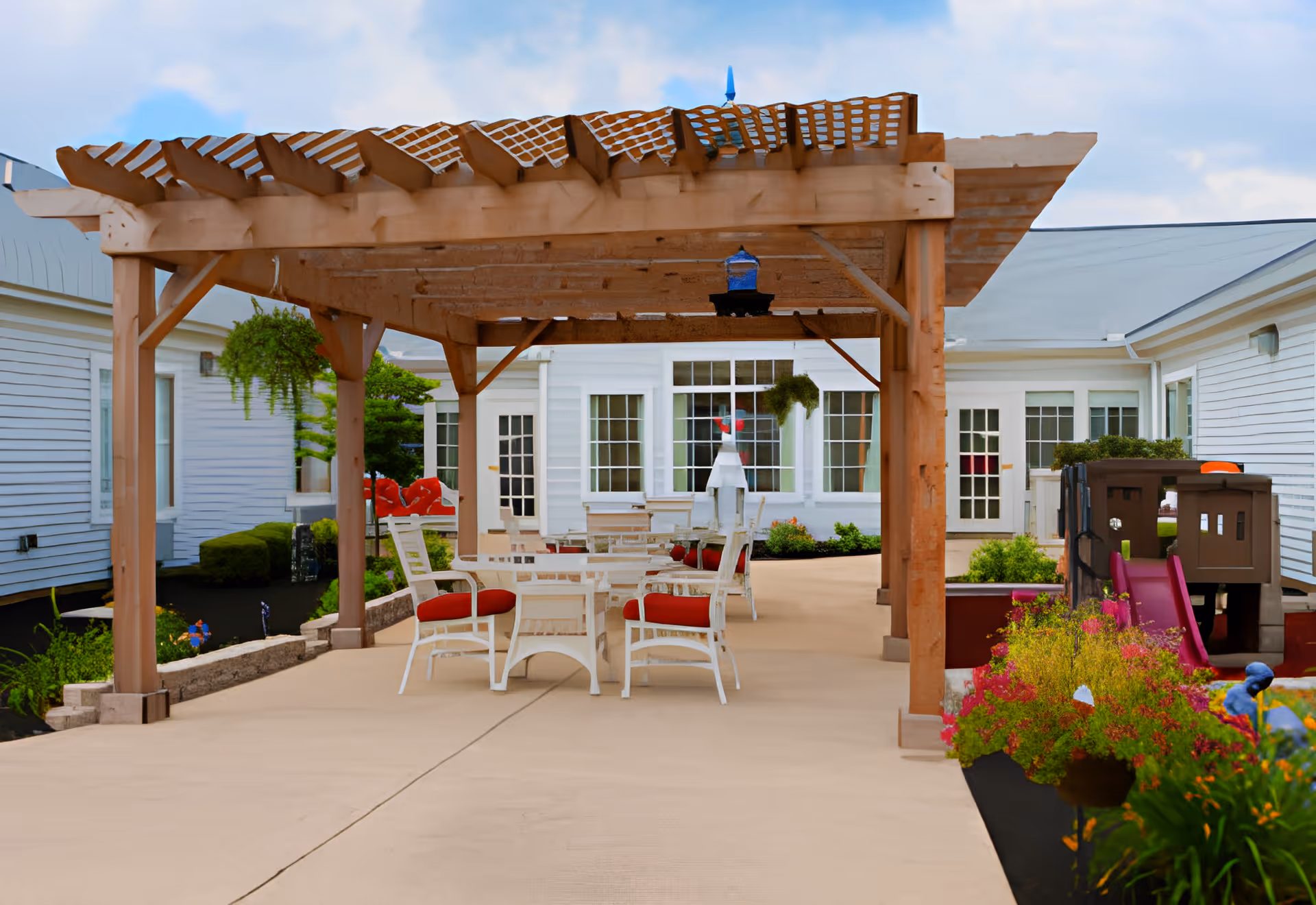 Outdoor patio area at Gardens At St Henry featuring a wooden pergola with hanging plants, white chairs with red cushions around tables, colorful flower beds, and a children's play structure with a slide. The background shows white building walls with multiple windows and doors.