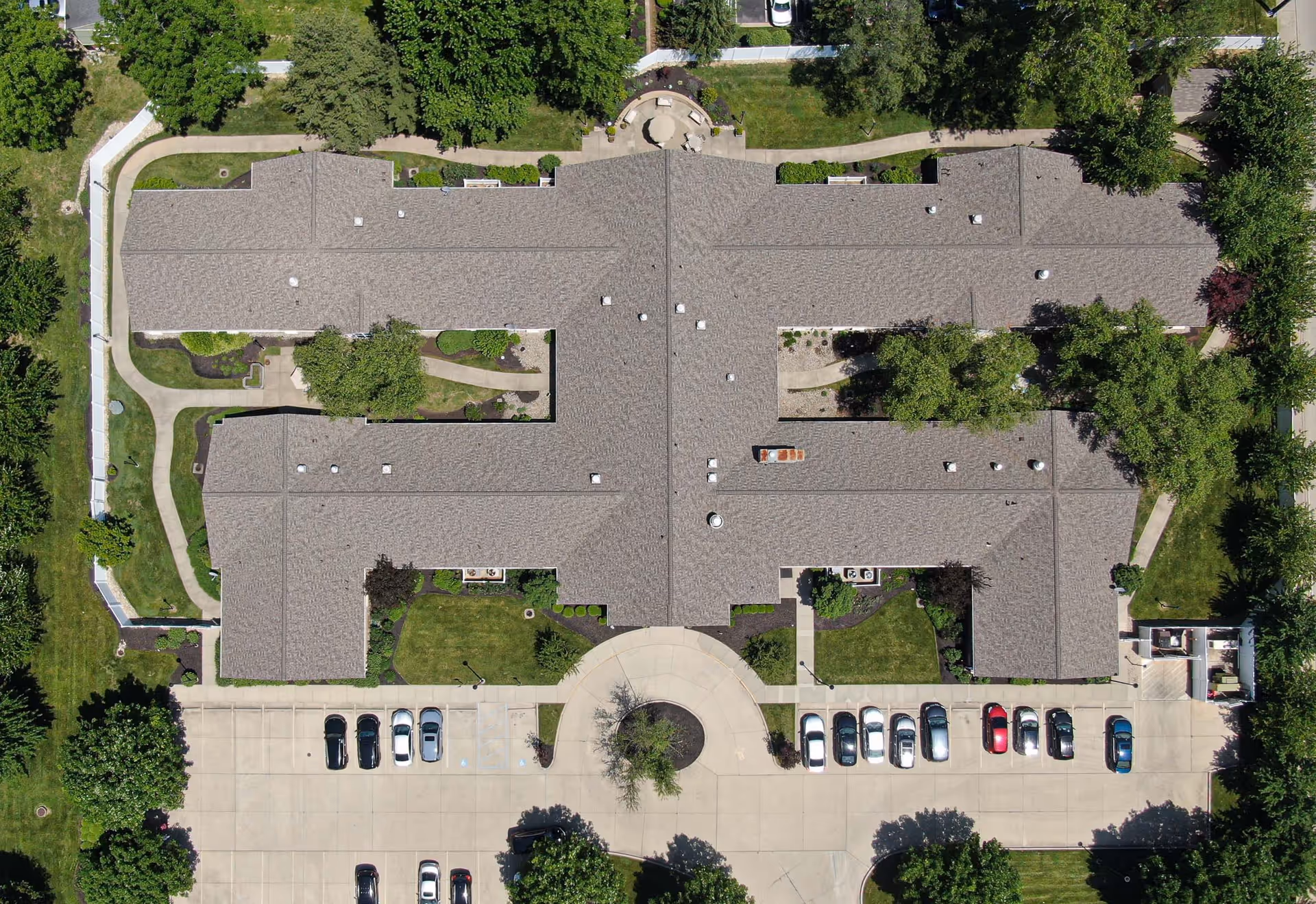 Aerial view of Arden Courts - ProMedica Memory Care Community (Kenwood) showing the building's roof, surrounding greenery, pathways, and a parking lot with several cars.