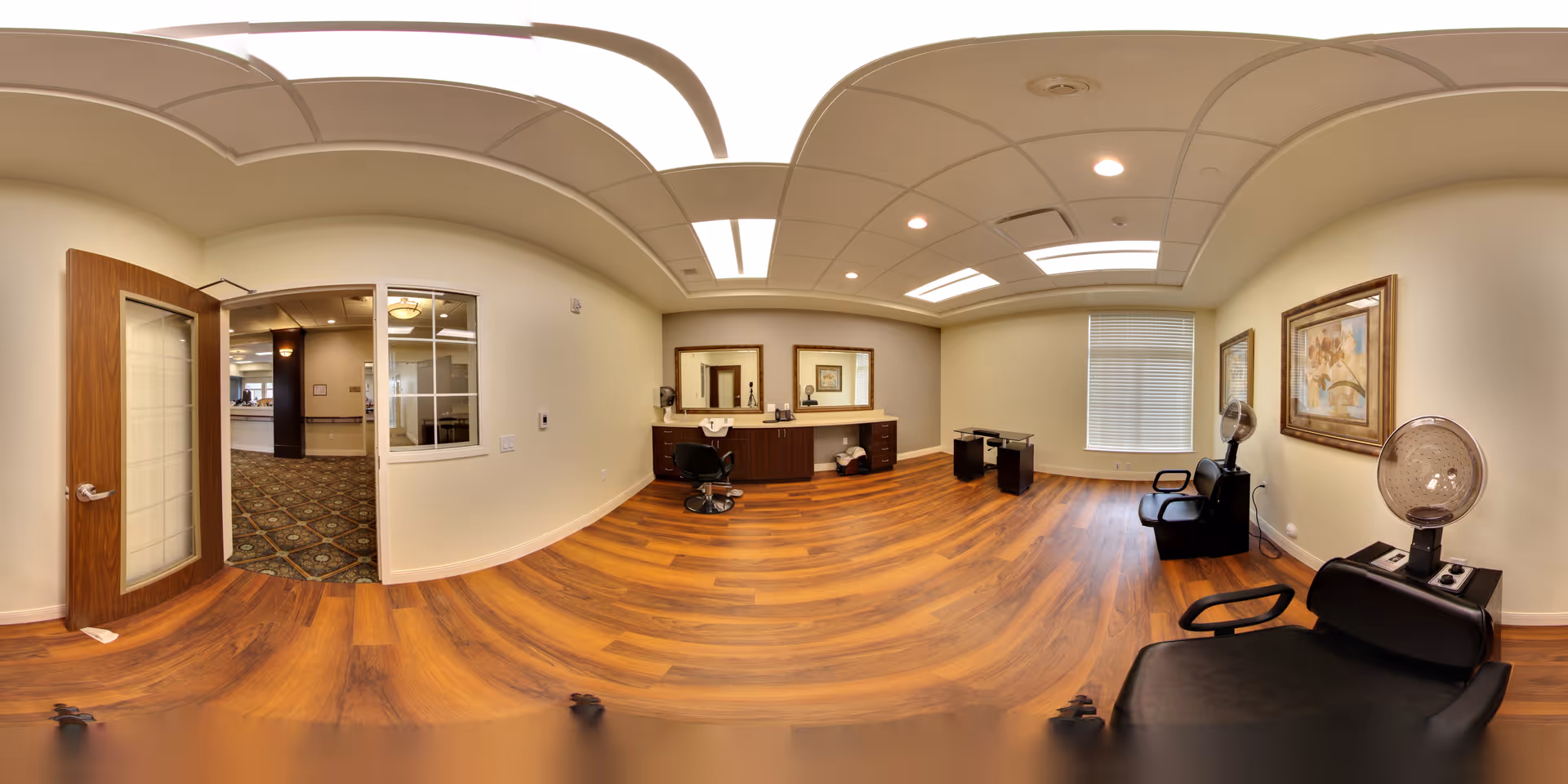 Interior view of a hair salon area within a senior living facility. The room features wooden flooring, two black salon chairs with hair dryers, a counter with two mirrors, and a window with blinds. The walls are painted light beige and decorated with framed artwork. A door with frosted glass panels opens to a carpeted hallway.
