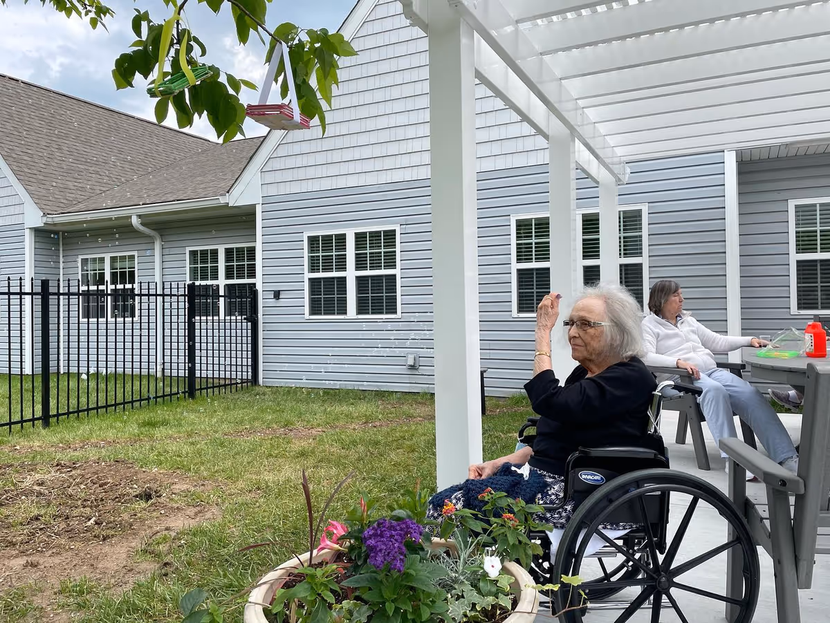 An elderly woman in a wheelchair blowing bubbles outside under a white pergola, with another woman sitting at a table in the background. The scene is set in a grassy courtyard area with a gray building and black metal fence surrounding it.