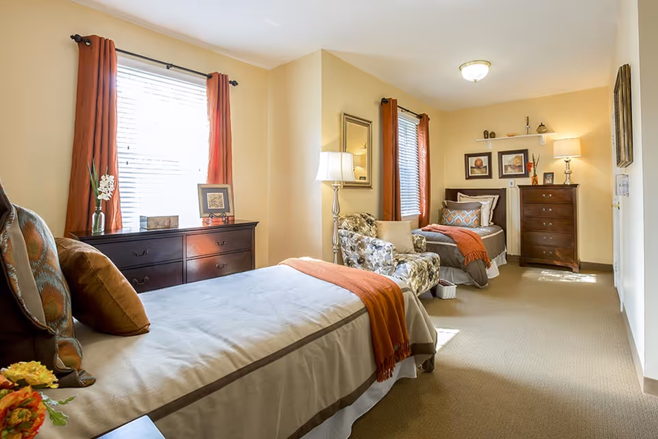 A bright and cozy bedroom with two single beds, each with pillows and orange throws. The room features two windows with orange curtains, a floral armchair, a floor lamp, two dark wooden dressers, framed artwork on the walls, and a beige carpeted floor.