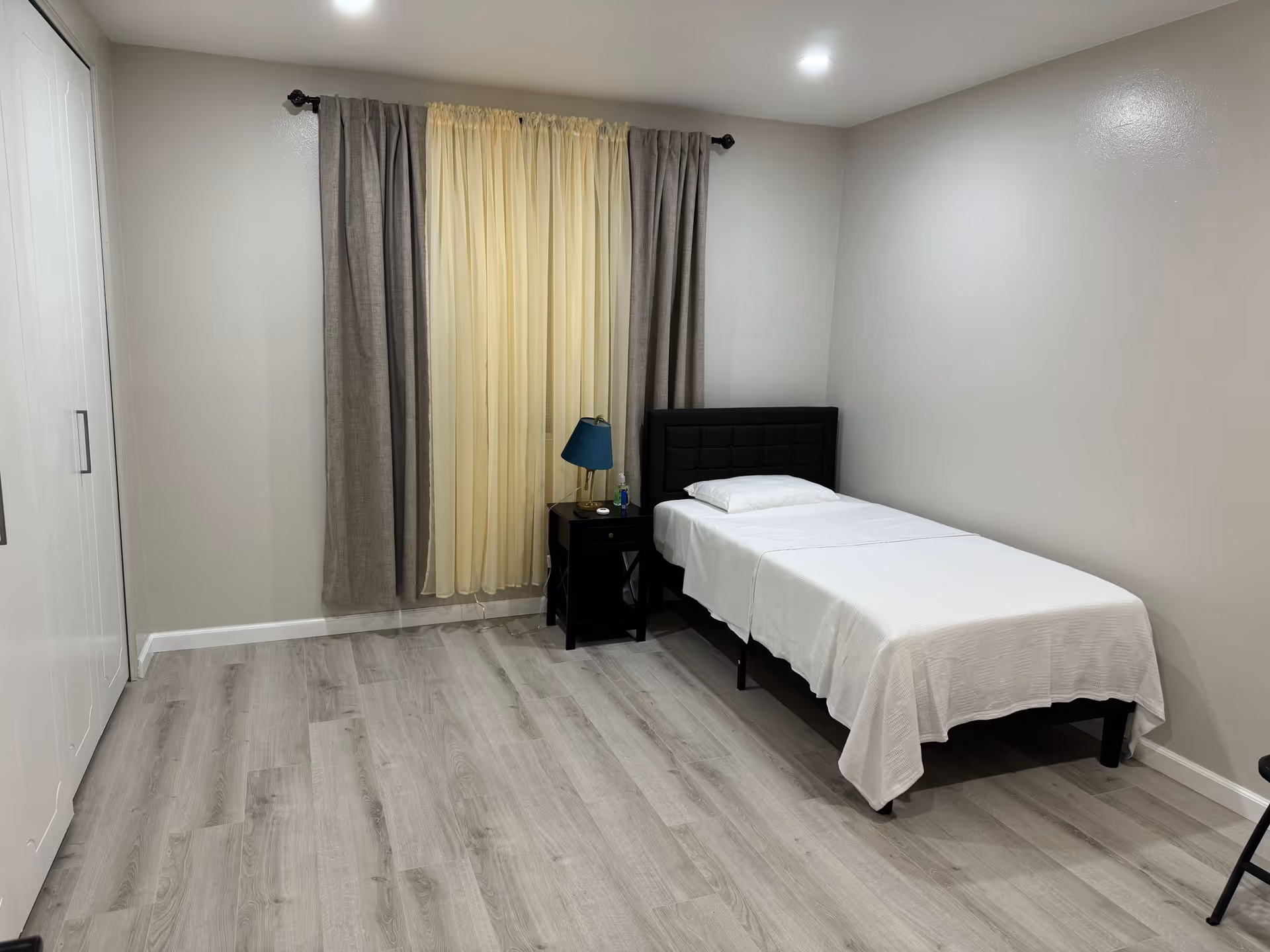 Simple single-occupancy bedroom with a twin bed, nightstand, lamp and curtained window.