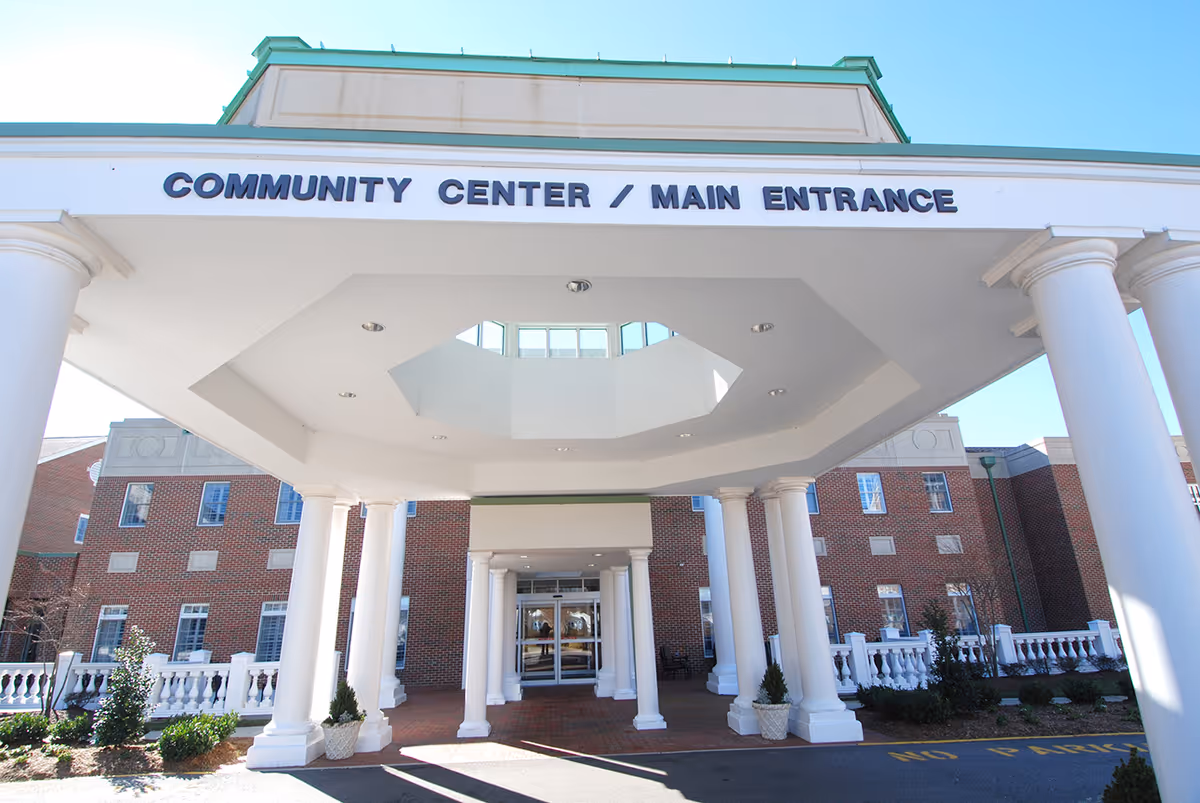 Entrance to a community center with white columns supporting a covered driveway. The sign above reads 'COMMUNITY CENTER / MAIN ENTRANCE'. The building is made of red brick with multiple windows and some landscaping with bushes and small trees.