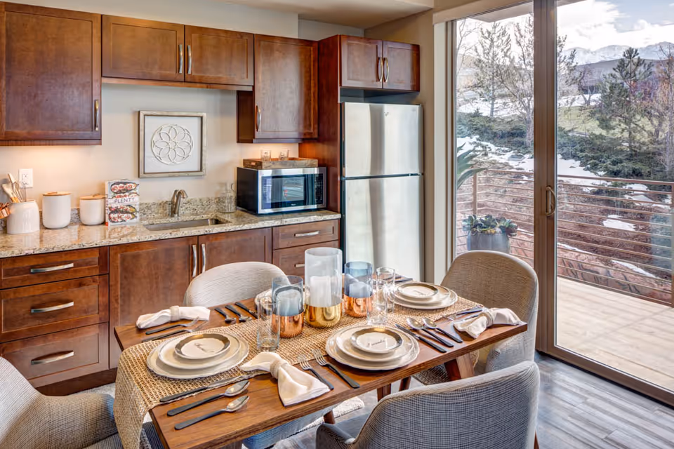 A modern kitchen and dining area with wooden cabinets, granite countertops, a stainless steel refrigerator, and a microwave. The dining table is set with plates, cutlery, napkins, and decorative candles. Large sliding glass doors open to an outdoor view with trees and mountains.
