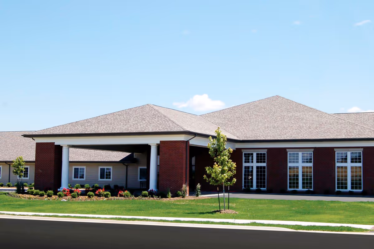 Exterior view of a single-story senior living facility building with a covered entrance supported by white columns, red brick walls, large windows, and a well-maintained lawn with small trees and flower beds under a clear blue sky.