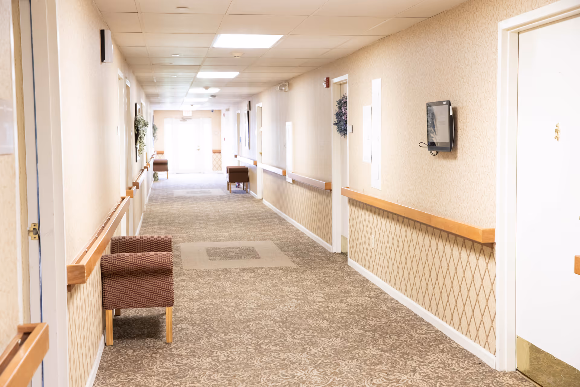 A long, well-lit hallway in a senior living facility with beige patterned carpet and wallpaper. Several doors line the walls, some decorated with wreaths. There are wooden handrails along both sides of the hallway and a few upholstered chairs placed against the walls. The hallway ends with double doors letting in natural light.