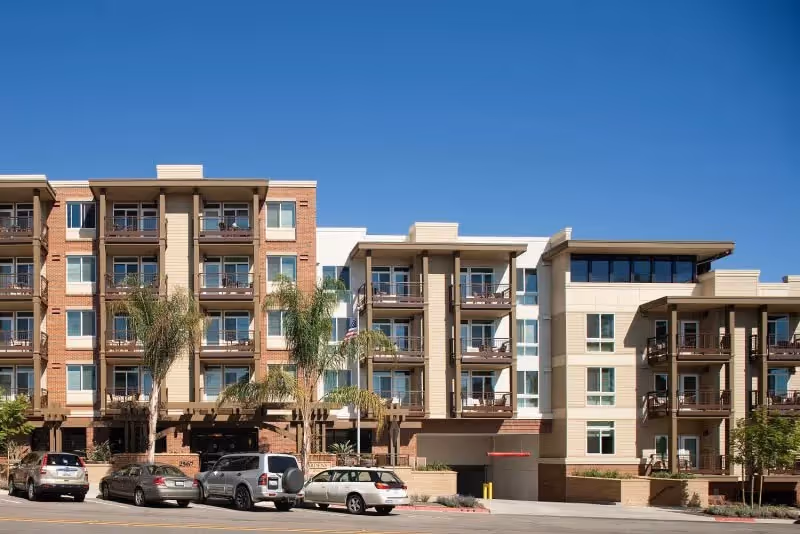 Street-level view of a multi-story brick and stucco residential building with balconies, palm trees, and cars parked in front under a clear blue sky.