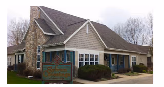 Exterior view of a single-story senior living facility building with beige siding, a stone chimney, multiple windows, and a covered entrance. There are bushes and a sidewalk in front, and a sign for the facility is visible near the entrance.