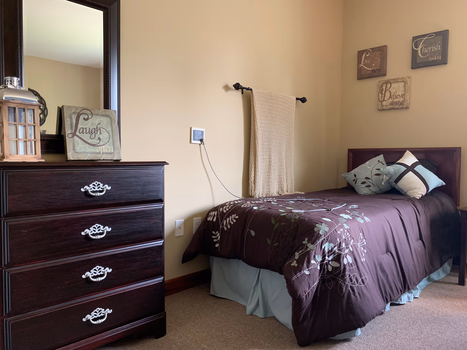 Small furnished bedroom with a single bed, dark wood dresser and mirror, decorative pillows and wall art.