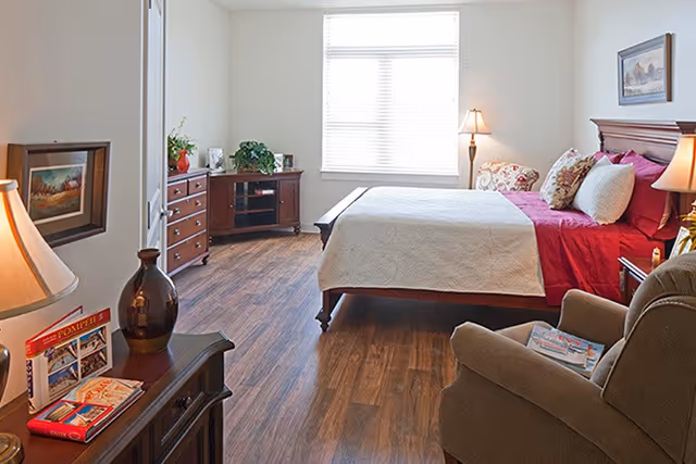 A bright bedroom with wooden flooring featuring a large bed with red and white bedding, a cushioned armchair with magazines, a wooden dresser with plants and picture frames, a window with blinds, and two table lamps providing warm lighting.