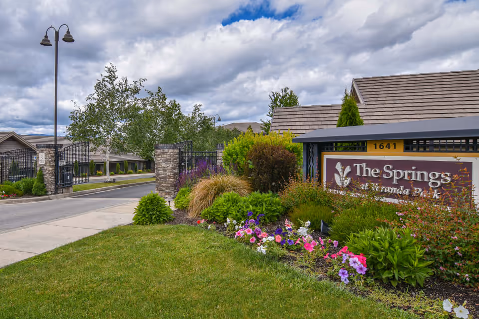 Entrance to The Springs at Veranda Park senior living facility featuring a landscaped garden with colorful flowers and shrubs, a stone and metal gated driveway, and a sign displaying the facility's name and address under a cloudy sky.