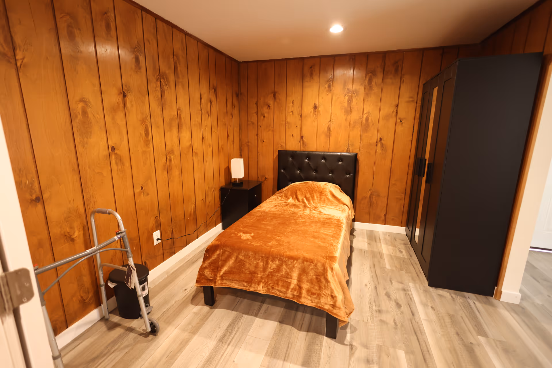 A small bedroom with wood-paneled walls, a single bed with a brown blanket and black tufted headboard, a black nightstand with a small lamp, a black wardrobe, and a walker near the door.