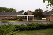 Front entrance of a single-story brick senior living facility with a covered drop-off, circular driveway, and landscaped shrubs.