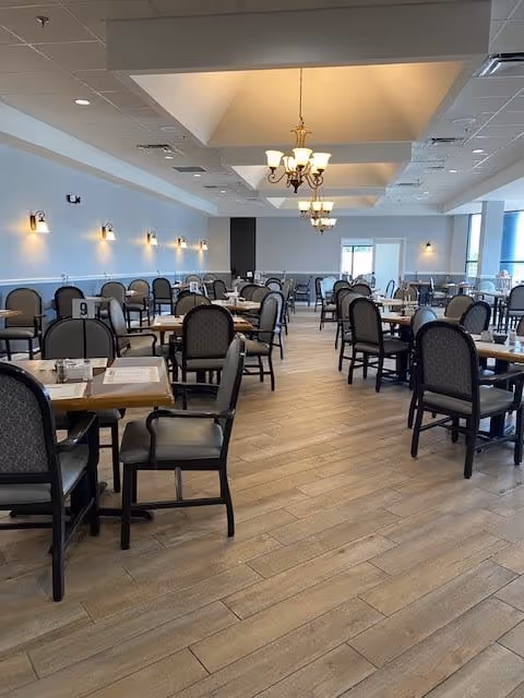 Spacious dining room with multiple tables and chairs arranged neatly. The room features wooden flooring, soft wall lighting, and chandeliers hanging from a recessed ceiling. Large windows on one side allow natural light to brighten the space.