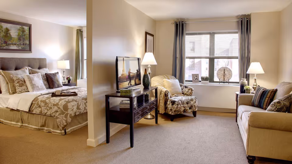 A cozy living space in a senior living facility featuring a bedroom area with a neatly made bed adorned with multiple pillows and a floral bedspread, separated by a partial wall from a living room area. The living room includes a patterned armchair, a beige sofa with decorative pillows, a TV on a dark wooden stand, two table lamps, and a window with blinds and curtains letting in natural light.