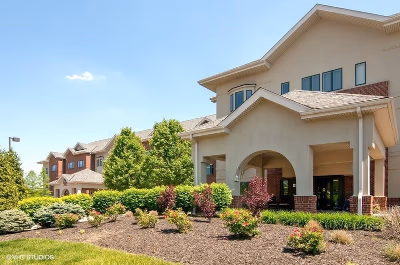 Exterior view of The Wellington at North Bend Crossing senior living facility showing a multi-story building with a covered entrance, surrounded by well-maintained landscaping including bushes, small trees, and flowers under a clear blue sky.