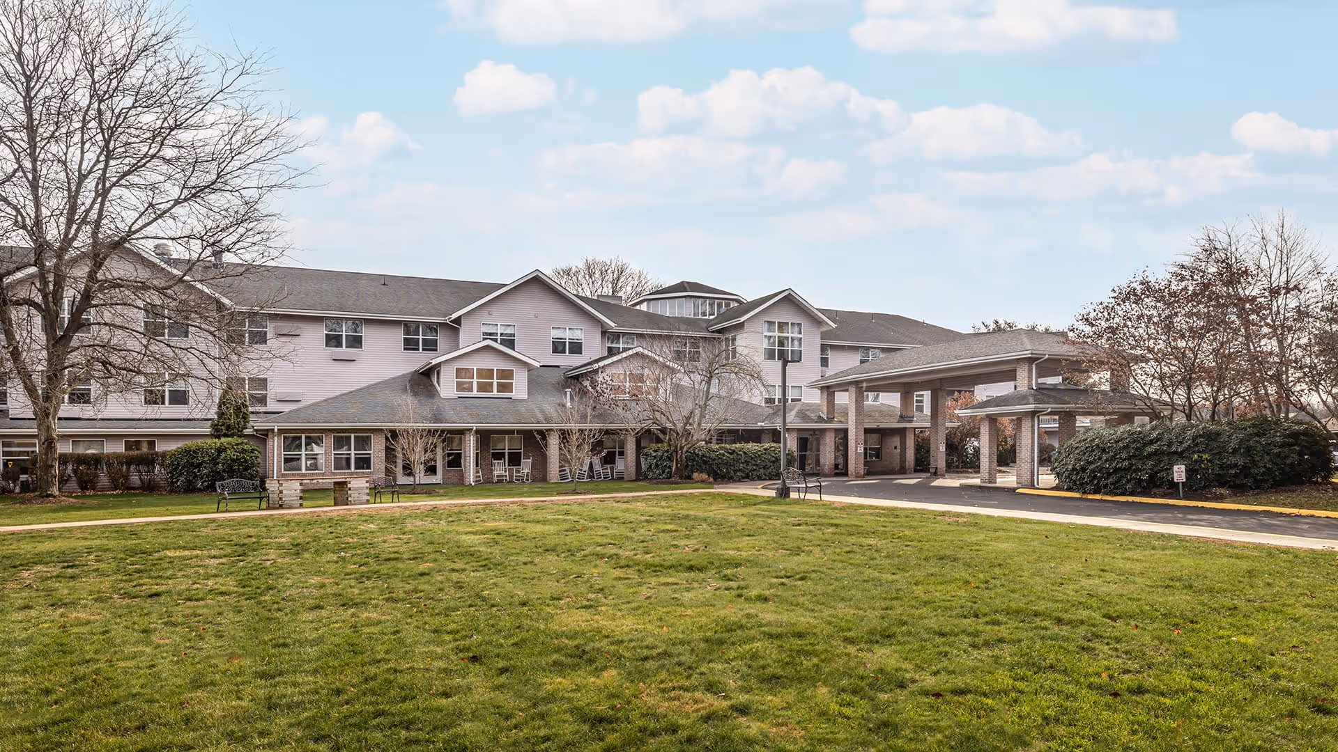 Exterior view of Holiday Quail Run Estates, a multi-story senior living facility with a covered entrance, surrounded by a grassy lawn and some leafless trees under a partly cloudy sky.