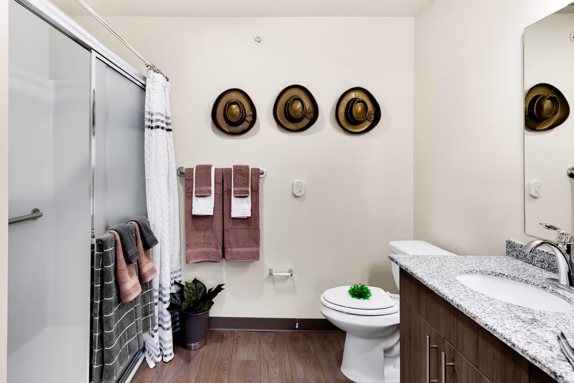 A clean bathroom with a shower enclosed by frosted glass sliding doors and a white shower curtain. There are several towels hanging on racks, including brown and gray towels. Three decorative hats are mounted on the wall above the towel rack. A white toilet with a green decorative bow on the lid is next to a granite countertop with a sink and faucet. A small potted plant is placed on the floor near the shower.