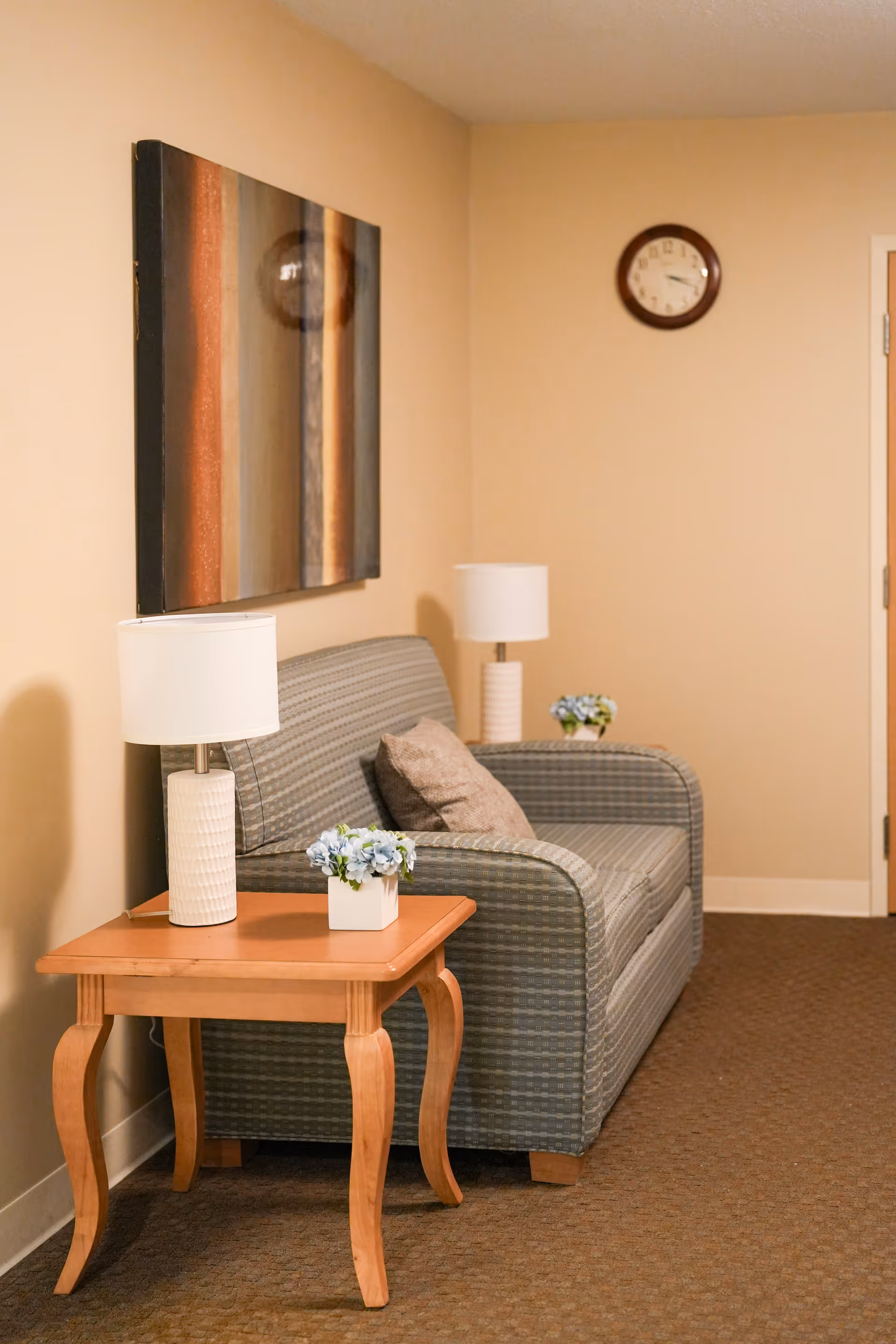 A cozy sitting area with a striped upholstered loveseat, a wooden side table with a white lamp and a small flower arrangement, a second white lamp and flower arrangement on the loveseat's armrest, a wall clock, and a modern abstract painting on the beige wall.