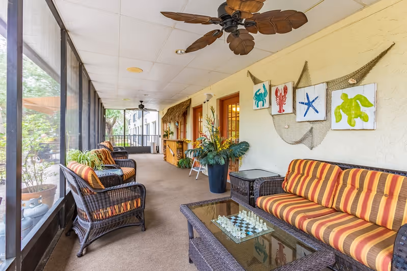 A screened-in porch area with wicker furniture including a striped cushioned sofa and chairs, a glass coffee table with a chess set, ceiling fans, potted plants, and wall decorations featuring sea creatures and a fishing net.