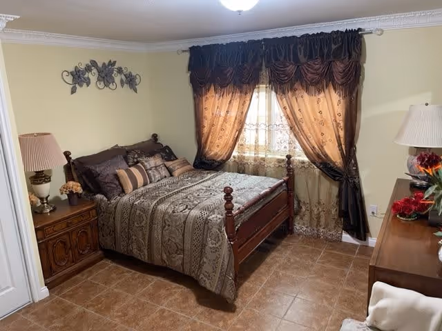 A cozy bedroom with a wooden bed frame and a patterned bedspread in shades of brown and beige. The bed is adorned with multiple decorative pillows. There is a wooden nightstand with a lamp and a small flower arrangement on the left side of the bed. A window with sheer and dark brown curtains is centered on the wall behind the bed. On the right side, there is a wooden dresser with a lamp and a vase of red flowers. The floor is tiled with brown tiles, and the walls are painted a light yellow color.