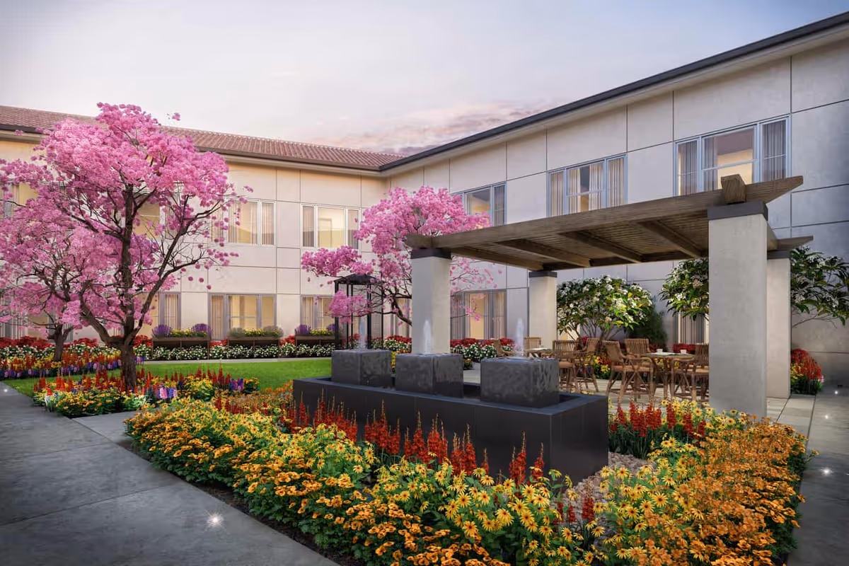 A landscaped outdoor courtyard with pink flowering trees, colorful flower beds, a pergola-covered seating area and a central water feature in front of a two-story building.