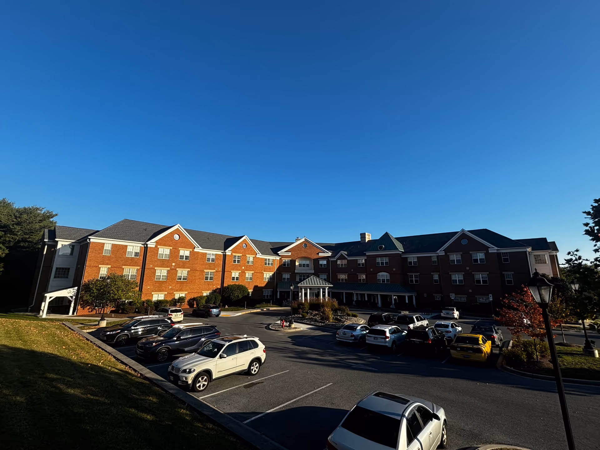 Exterior view of a large three-story brick building with multiple windows and a covered entrance. Several cars are parked in the parking lot in front of the building under a clear blue sky.