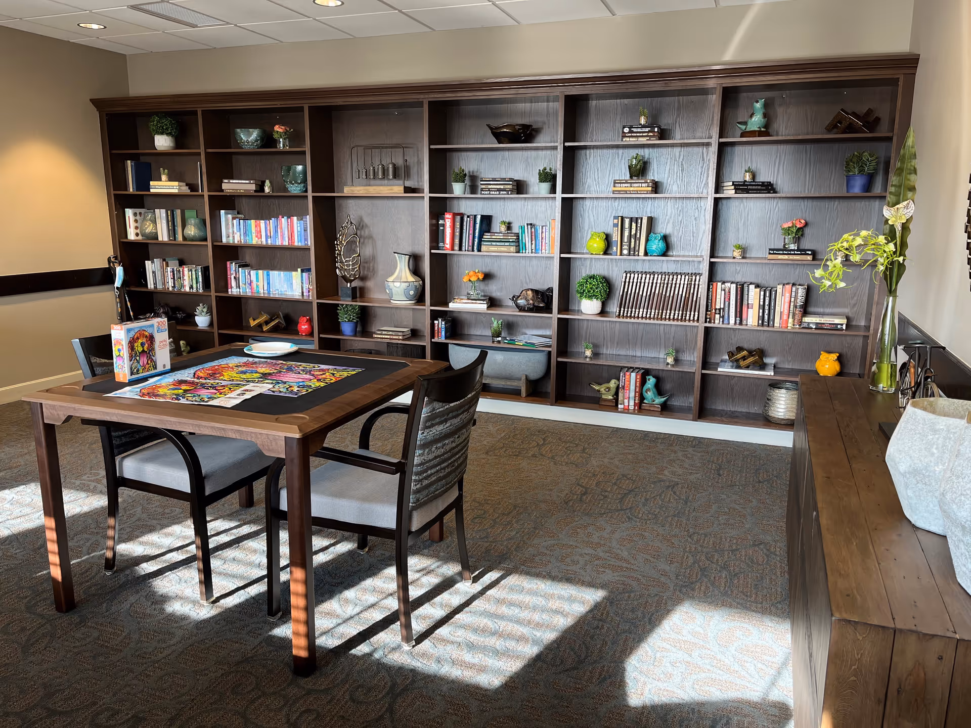 A cozy room with a large wooden bookshelf filled with books, decorative items, and small plants. In front of the bookshelf is a wooden table with two chairs, a partially completed colorful puzzle, and a puzzle box on the table. The room has carpeted flooring and soft natural light coming through a window.