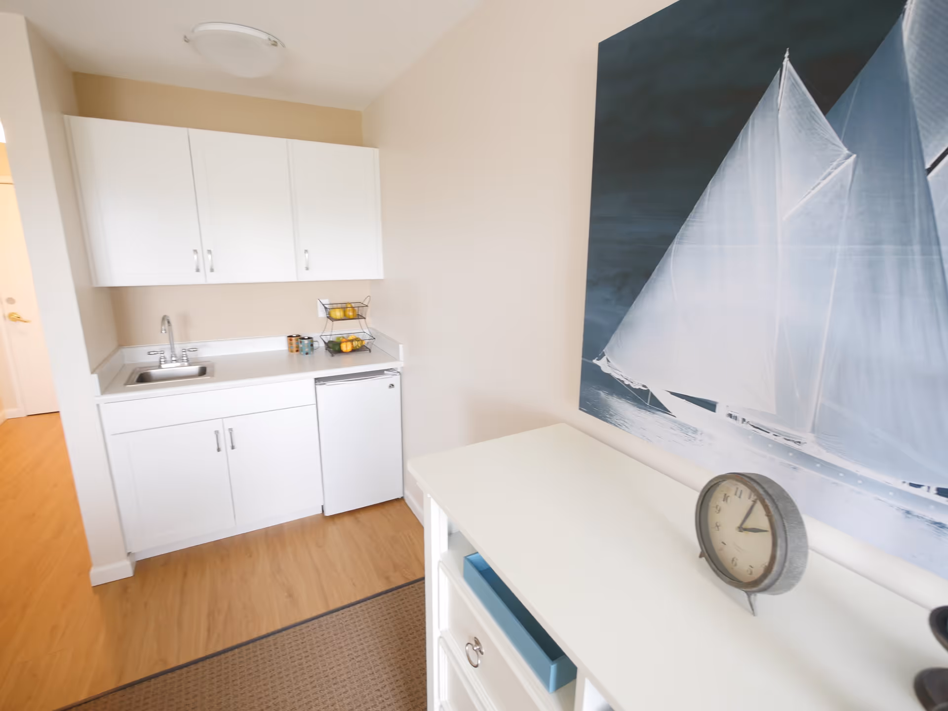 Small kitchenette area with white cabinets, a sink, a mini fridge, and a countertop holding a fruit basket and mugs. Adjacent to the kitchenette is a white dresser with a round clock on top and a large wall art of a sailboat in the background.