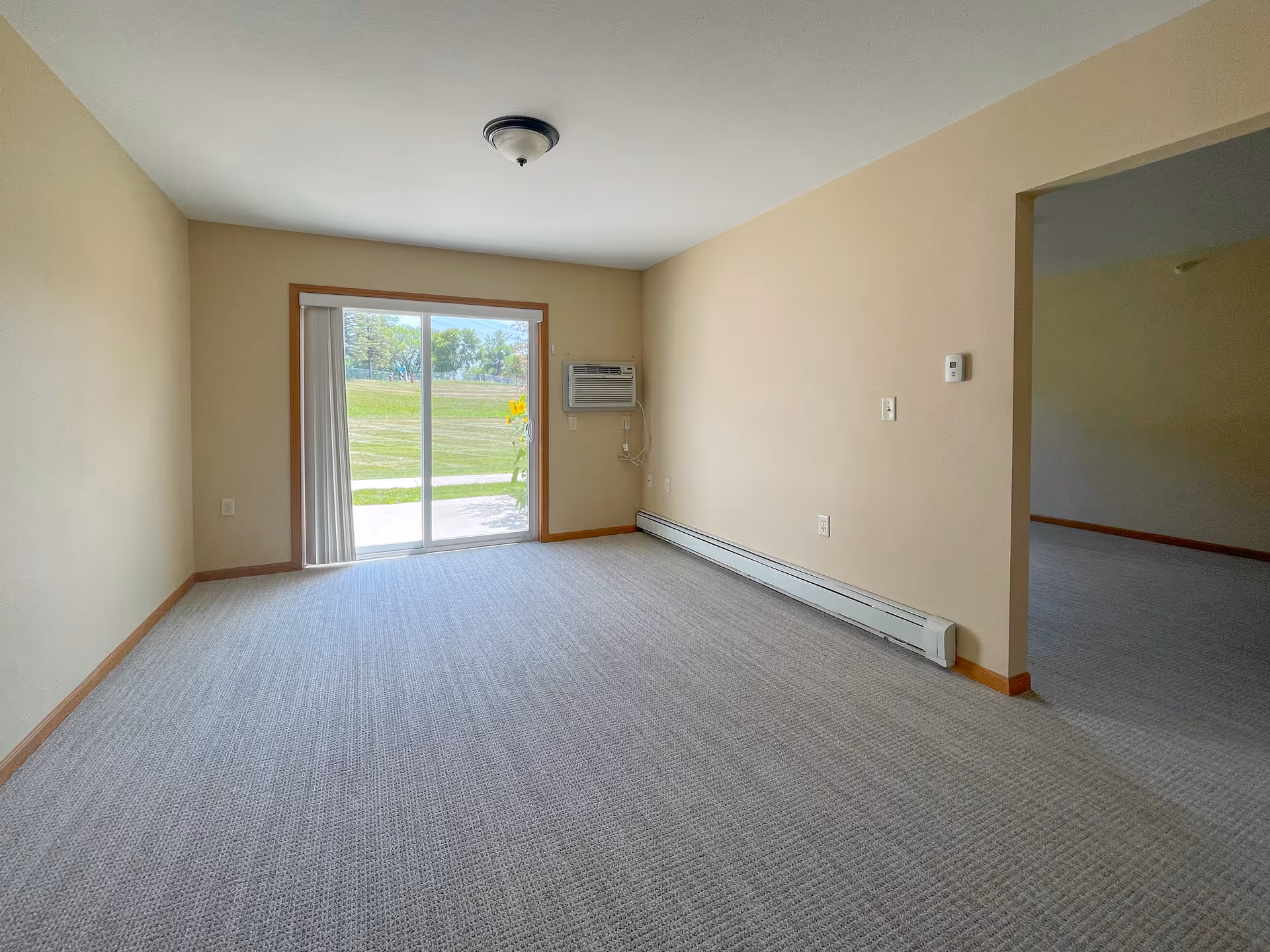 Empty room with beige walls and carpeted floor, featuring a sliding glass door leading outside to a grassy area. There is a wall-mounted air conditioning unit on the right wall and a ceiling light fixture in the center of the ceiling. An open doorway on the right side leads to another room.