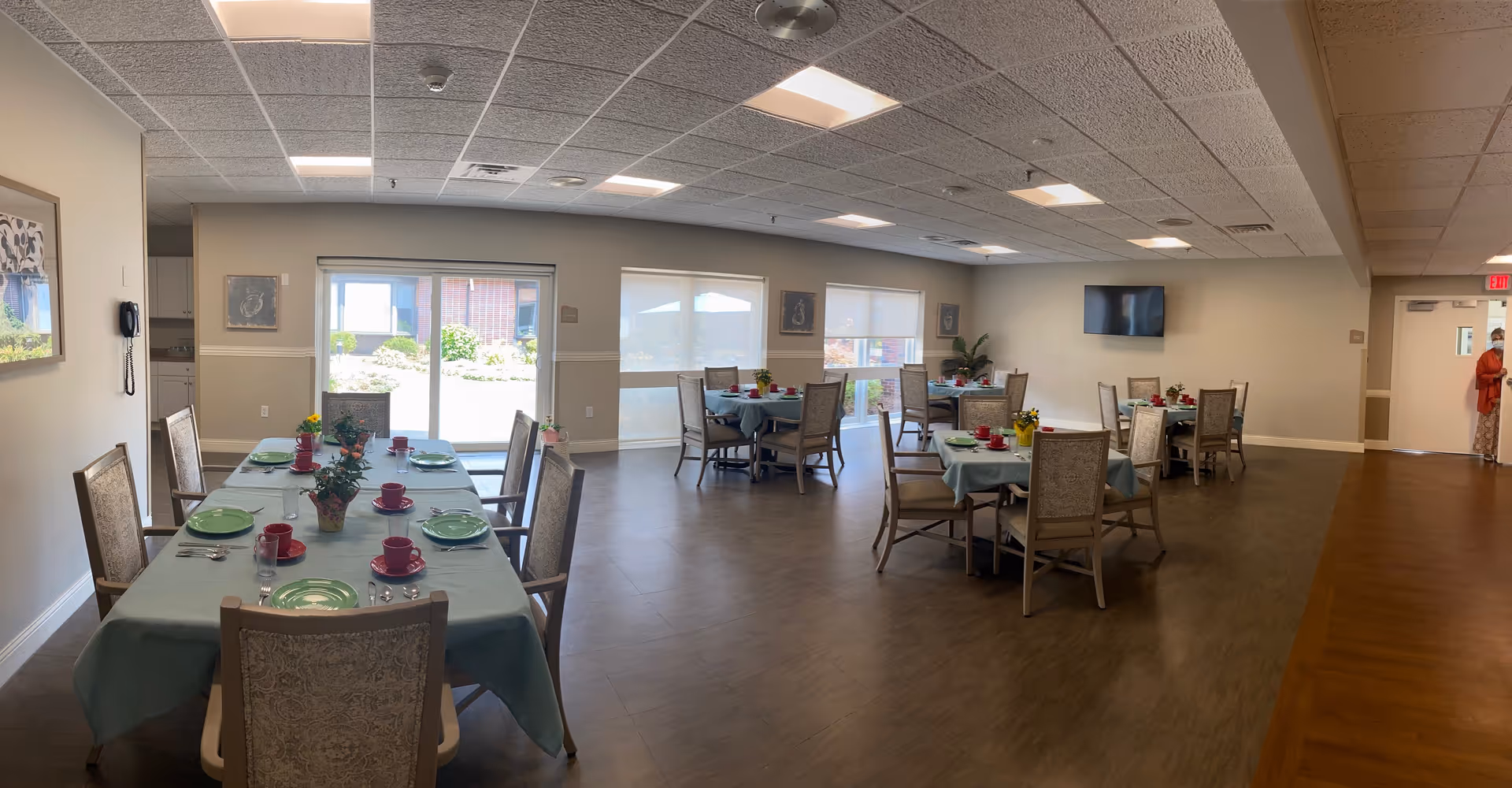 Spacious communal dining room with multiple tables set with plates, cups and centerpieces, chairs, windows and a wall-mounted TV.