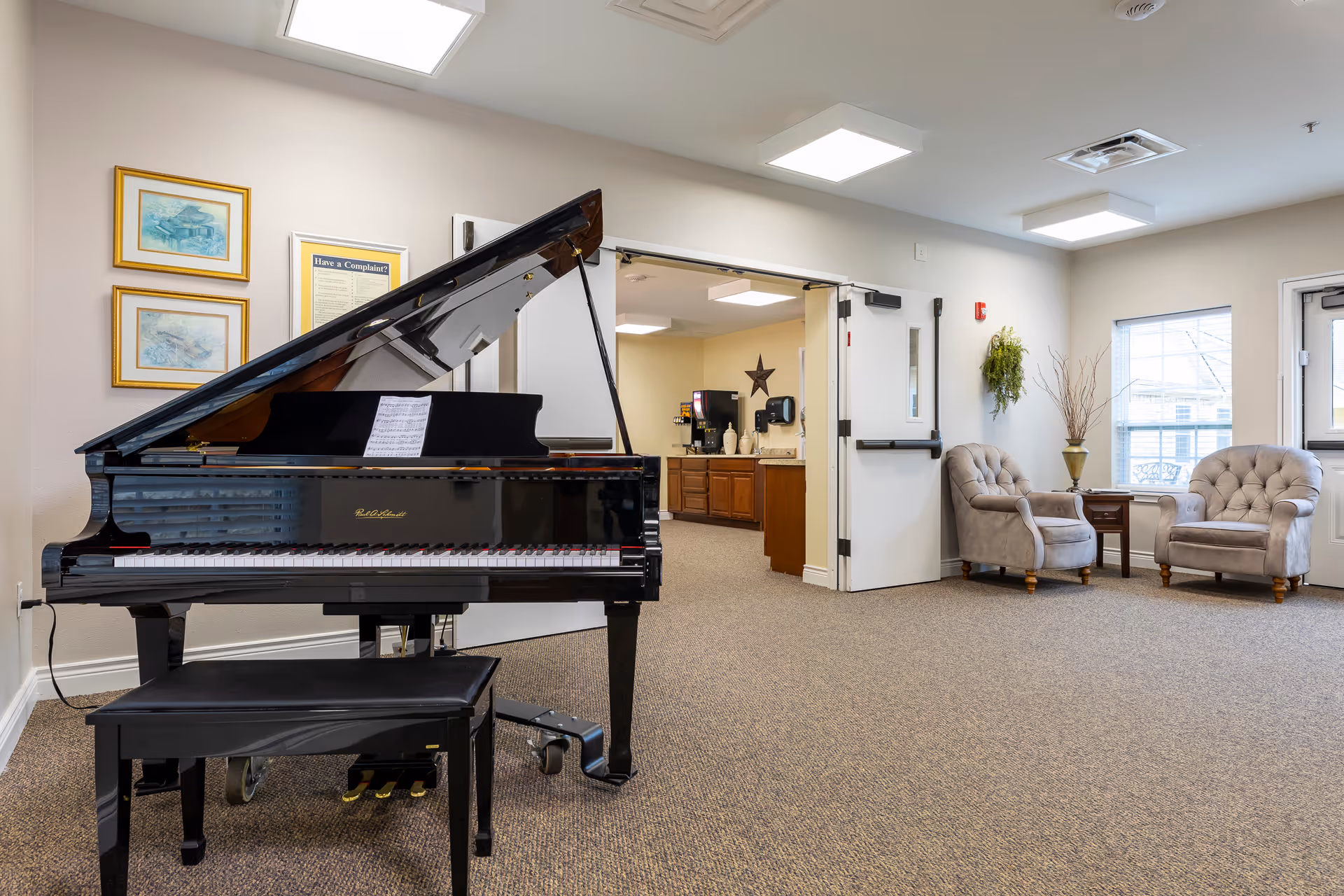 A bright room featuring a black grand piano with sheet music on the stand and a matching piano bench. In the background, there is an open doorway leading to a kitchenette area with wooden cabinets and a coffee machine. To the right, two tufted armchairs flank a small wooden table, with a window and a door letting in natural light. The walls are decorated with framed pictures and a hanging plant.