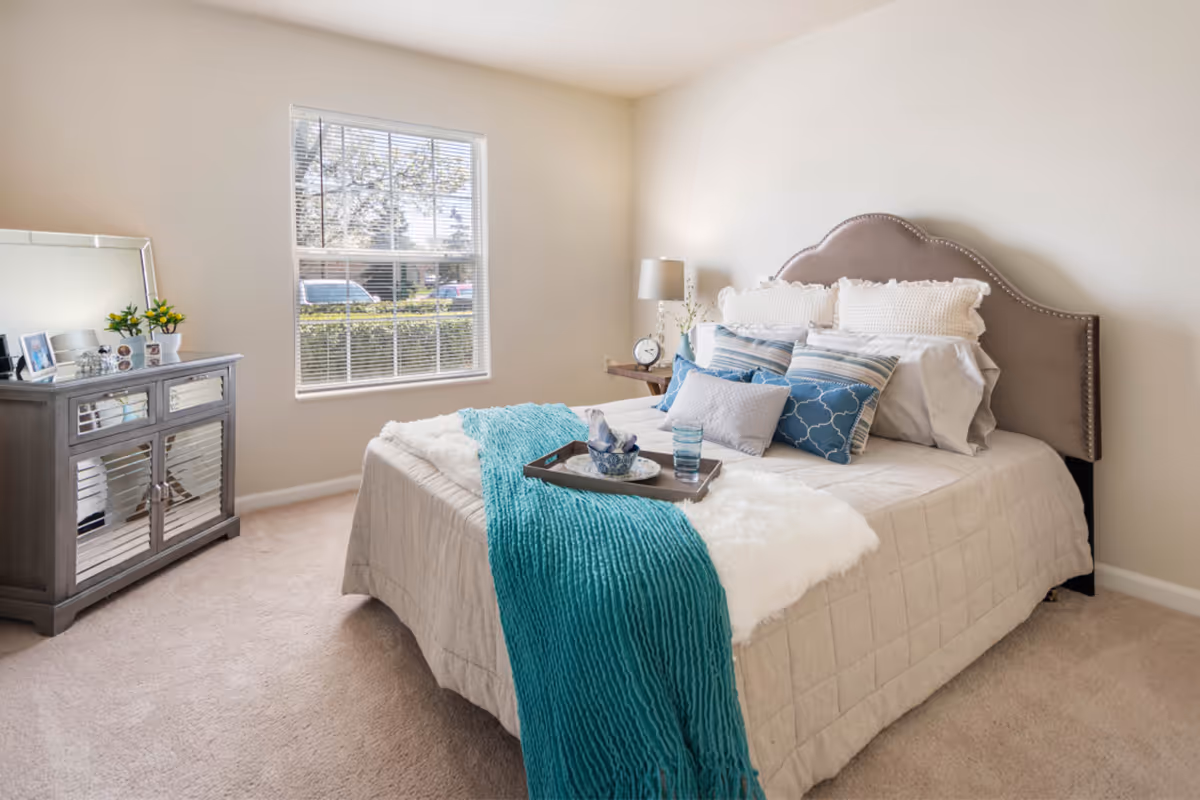 Bright bedroom with a neatly made bed featuring a teal throw and decorative pillows, a window, and a mirrored dresser.