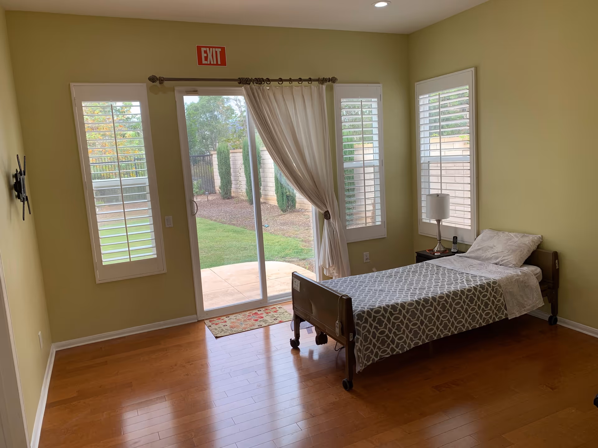 Single-occupancy bedroom with a hospital-style bed, nightstand and lamp, and a sliding glass door opening to a small patio.