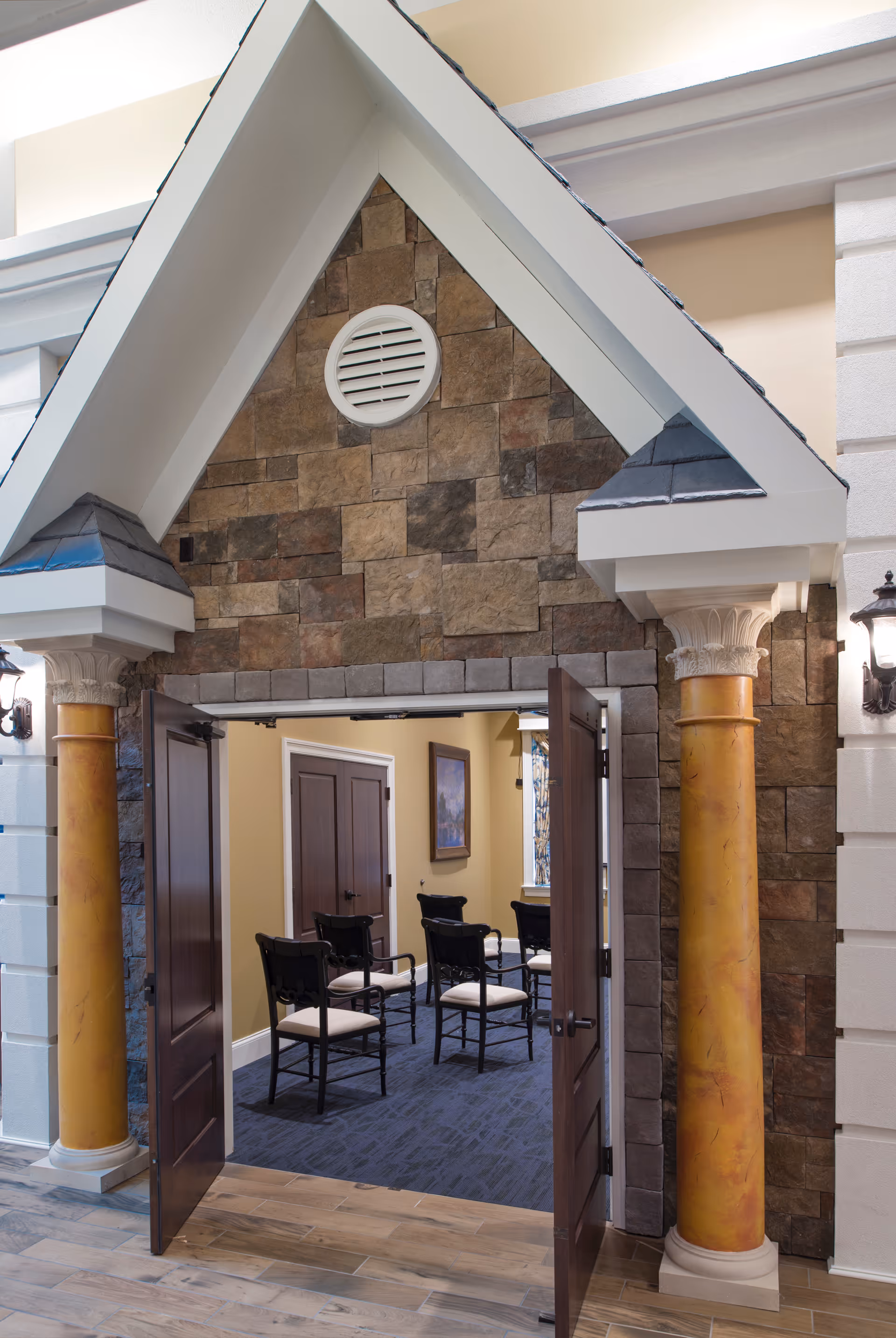 Entrance to a room with double wooden doors open, flanked by two decorative columns with yellow and white detailing. The walls around the entrance are made of stone tiles, and inside the room, there are several black chairs with white cushions arranged on a blue carpet. A framed picture hangs on the beige wall inside the room.