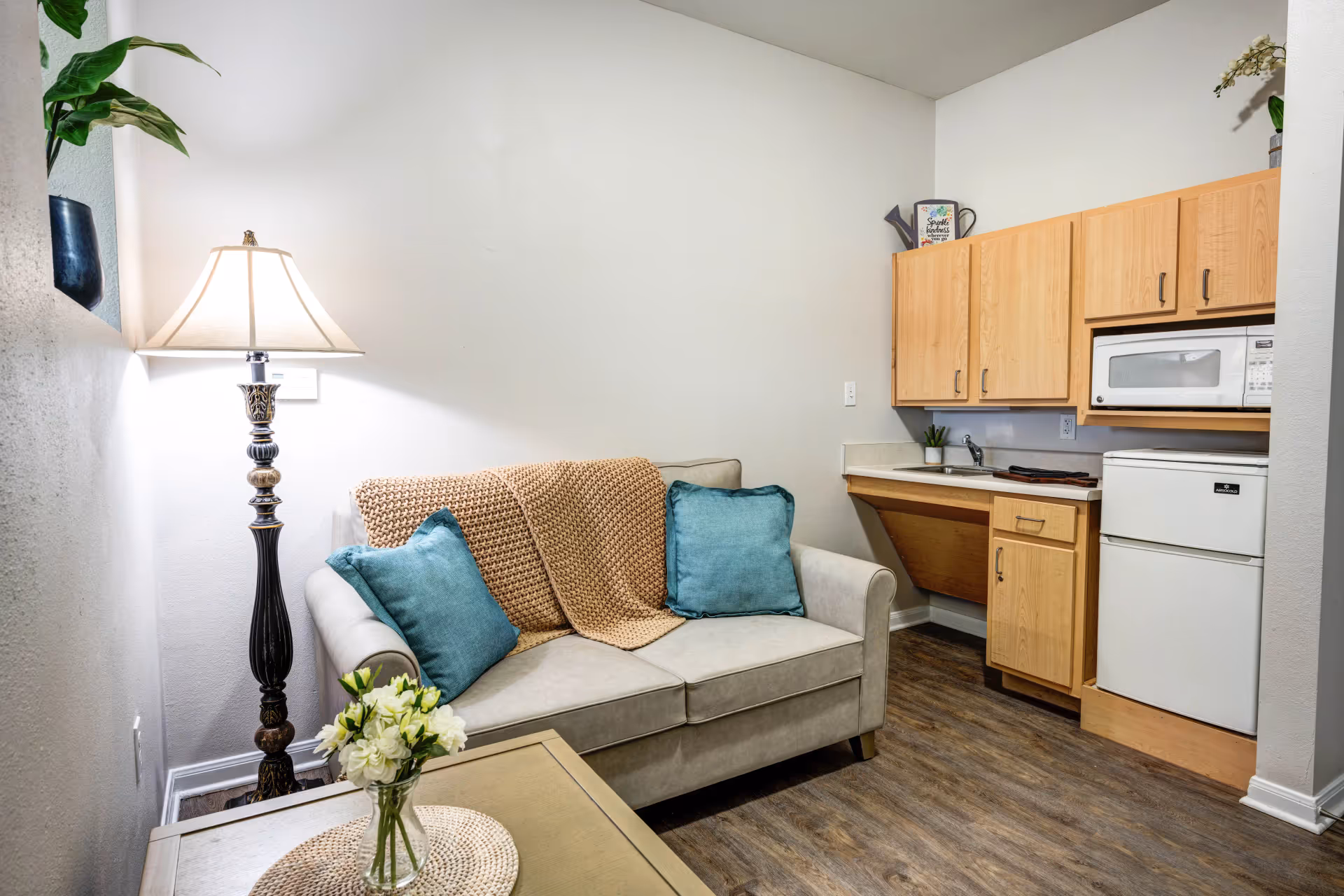 A small living area with a beige loveseat adorned with two teal pillows and a tan knitted throw blanket. Next to the loveseat is a tall decorative floor lamp and a small table with a vase of white flowers. In the background, there is a compact kitchenette with light wood cabinets, a white mini refrigerator, a microwave, and a small sink. The room has wood flooring and white walls with minimal decoration.