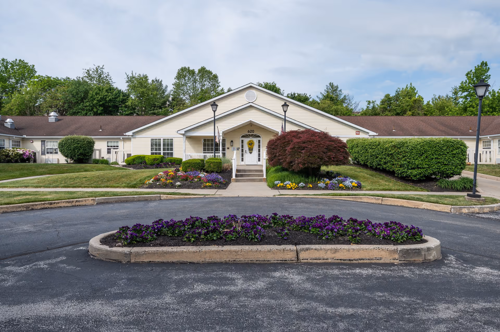 Front exterior view of a single-story building with beige siding and a brown roof, surrounded by well-maintained landscaping including bushes, colorful flowers, and a small tree. A circular driveway with a flower bed in the center is visible in the foreground. Two lampposts flank the entrance, which has steps leading up to a door with a wreath.