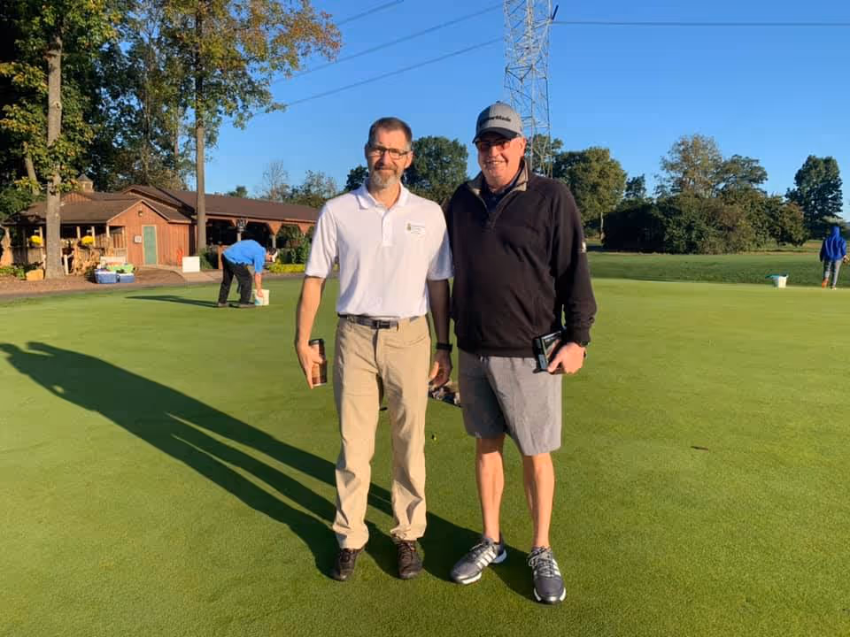 Two men standing on a well-maintained golf green with a building and trees in the background. One man is wearing a white polo shirt and beige pants, while the other is wearing a black jacket, gray shorts, and a cap. Another person is seen bending down near a bucket on the green, and another person is walking in the distance.