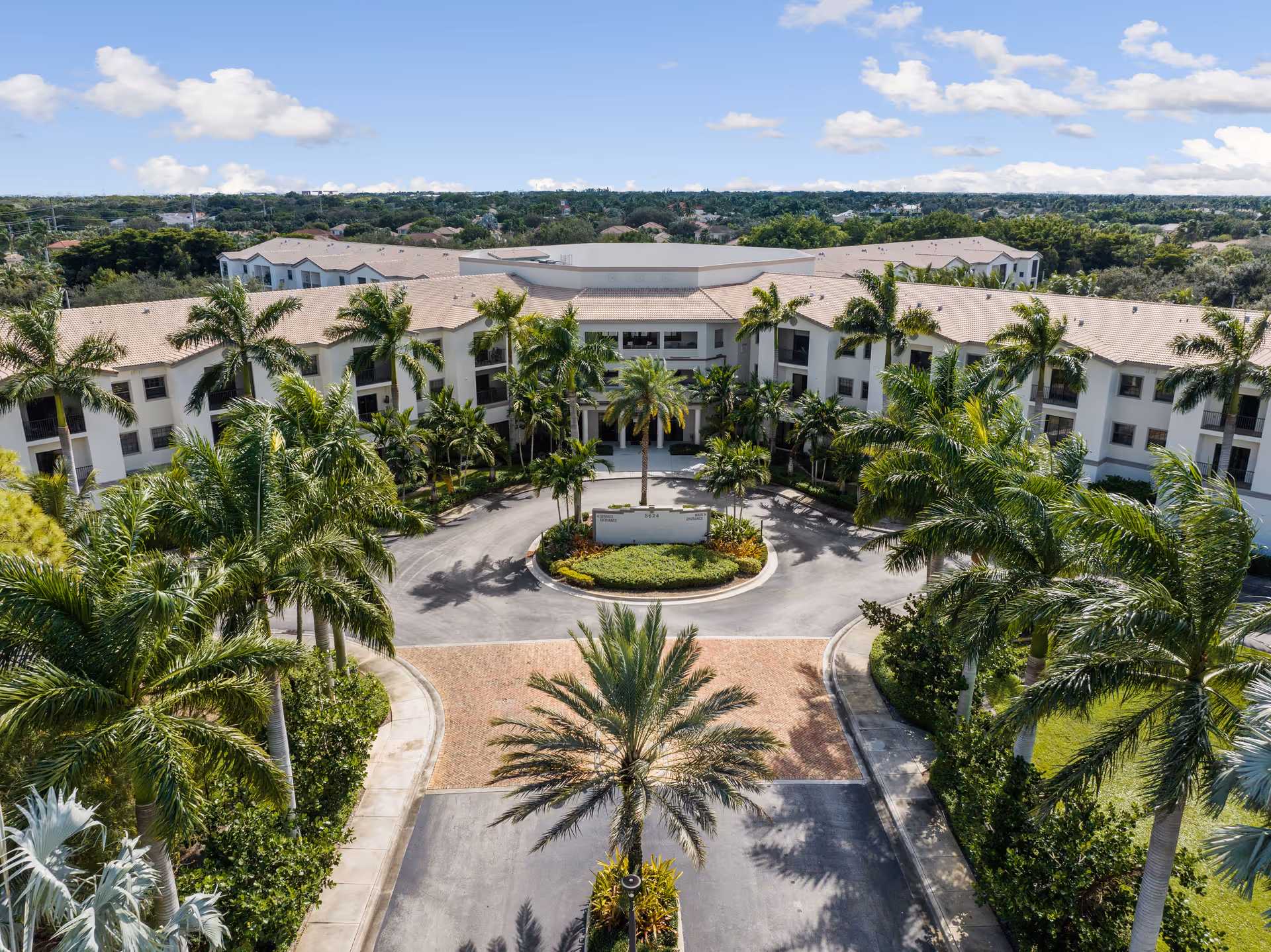 Aerial view of Verena at Delray senior living facility showing a large, U-shaped building surrounded by palm trees and greenery under a blue sky with scattered clouds. The entrance features a circular driveway with landscaped greenery in the center.