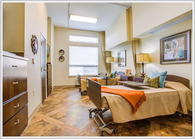 Bright, warmly decorated shared resident room with two adjustable beds, bedside tables, lamps, and a seating area.