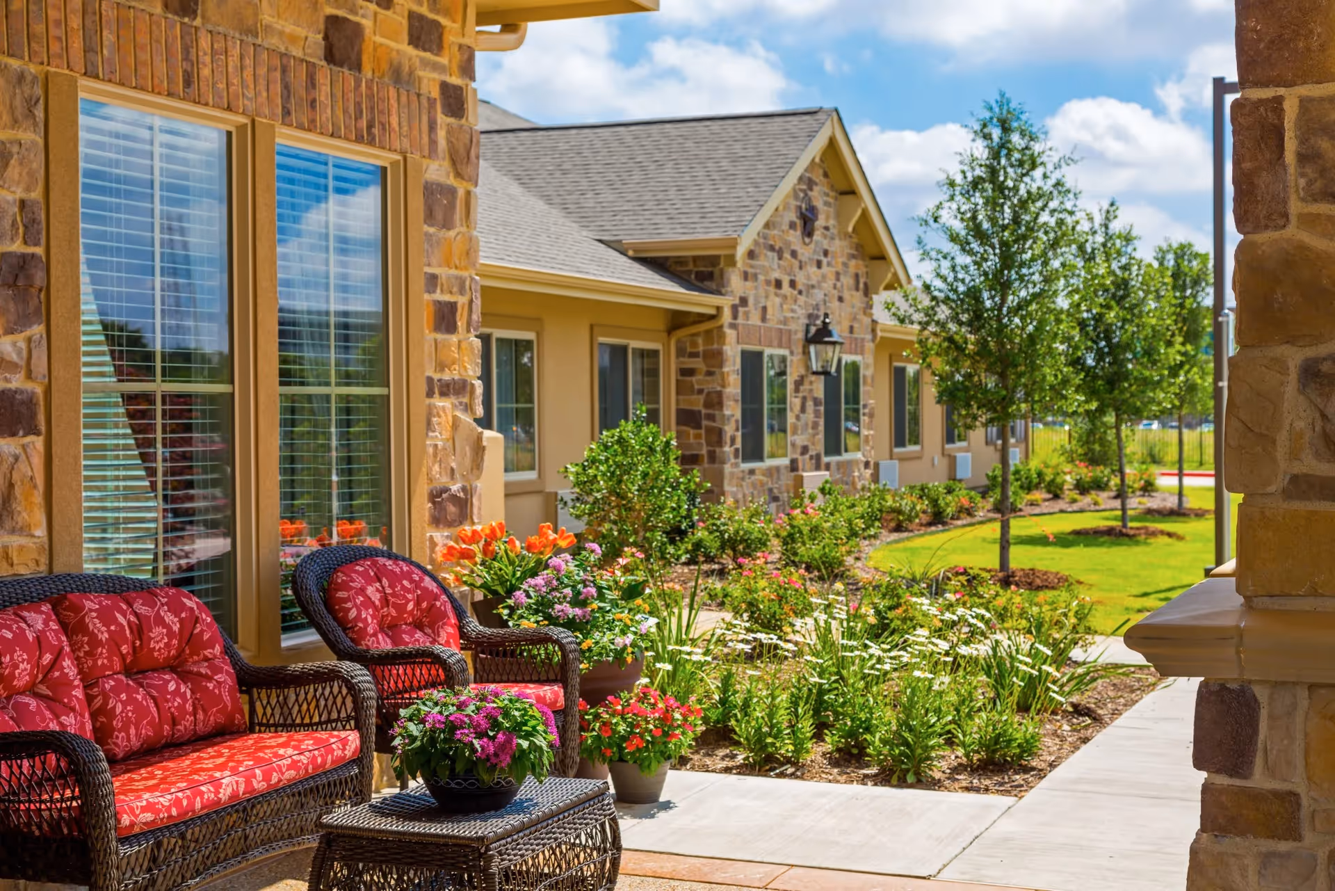 Outdoor patio area of a senior living facility with wicker furniture featuring red floral cushions, a small table with a potted plant, and a garden with various flowers and shrubs. The building has stone and beige siding with multiple windows and a walkway leading through a landscaped area with trees and grass under a partly cloudy sky.