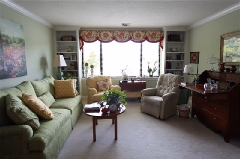 A cozy living room with a green sofa, two armchairs, a wooden coffee table with plants and decorative items, built-in bookshelves flanking a large window with floral valance, and a wooden desk with a lamp and decorative pieces.