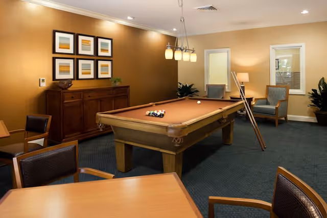 A cozy game room featuring a pool table with billiard balls arranged for a game. The room has warm beige walls, a wooden sideboard with framed artwork above it, several chairs around a wooden table in the foreground, and two armchairs with a floor lamp in the background. The room is softly lit with ceiling lights and a hanging light fixture above the pool table.