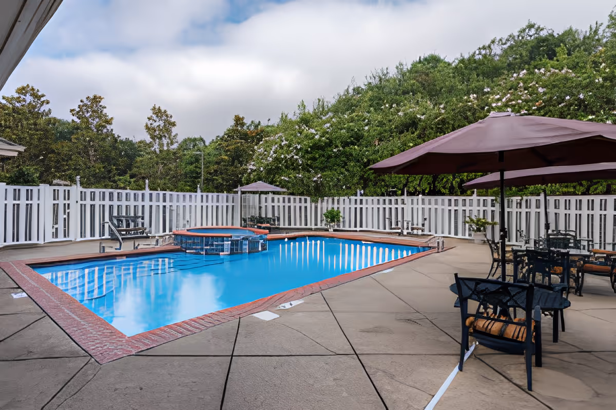 Outdoor swimming pool area with clear blue water, surrounded by a white fence and greenery. There are several black metal tables and chairs with brown umbrellas providing shade on the concrete pool deck.