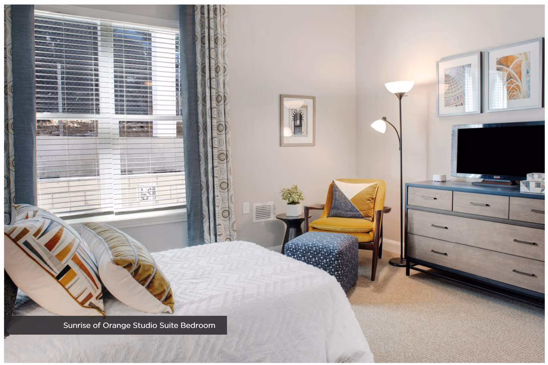 Bright studio bedroom with a bed, patterned pillows, window with blinds, a yellow armchair and ottoman, and a dresser with a TV.