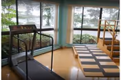 A small exercise room with a treadmill facing large windows that provide a view of trees and greenery outside. The room has a wooden floor and a padded exercise platform with steps on the right side.
