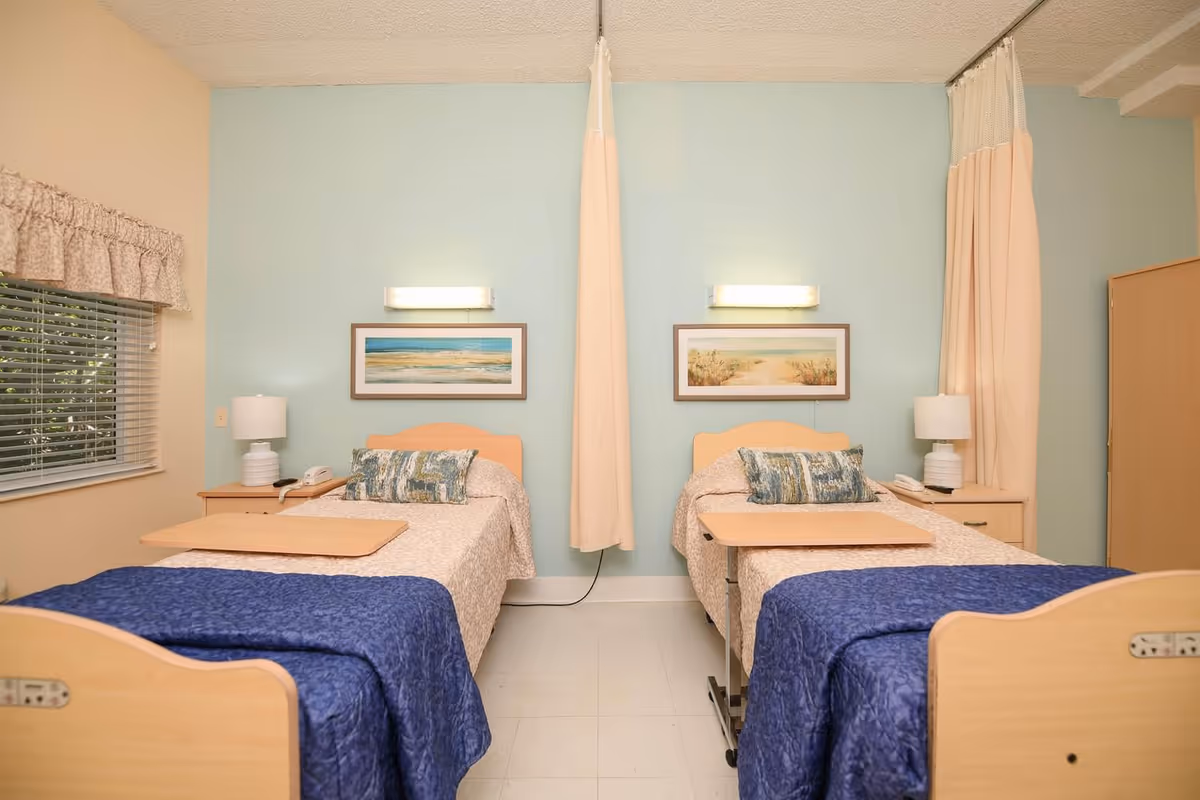 Two hospital-style twin beds with blue throws, bedside tables and lamps in a light-blue nursing facility room divided by a privacy curtain.