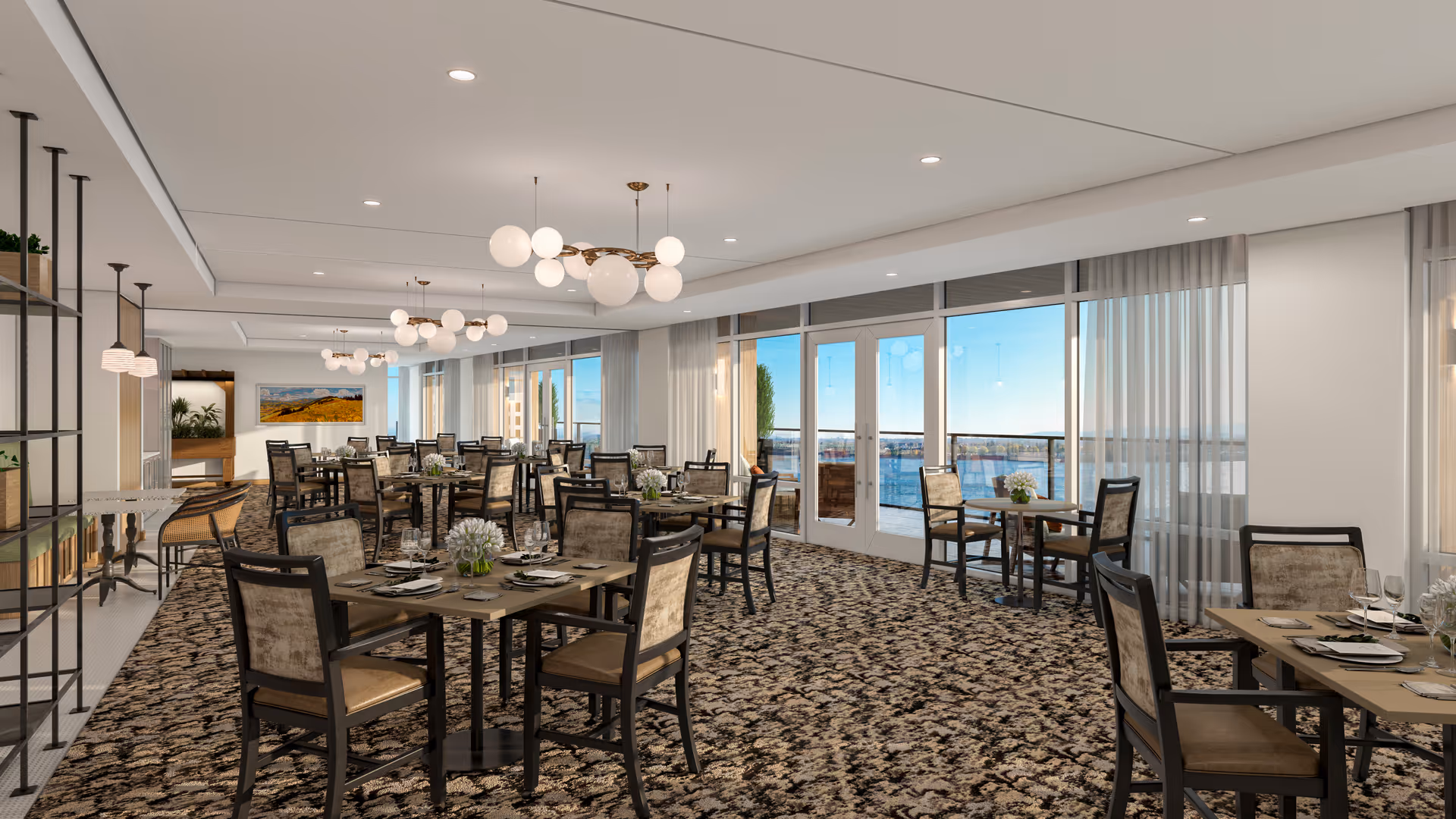 A spacious, well-lit dining room with multiple tables and chairs arranged neatly. The room features large windows and glass doors that offer a view of an outdoor waterfront area. Modern light fixtures hang from the ceiling, and the tables are set with plates, glasses, and small floral centerpieces. The carpet has a patterned design, and there are sheer curtains on the windows.