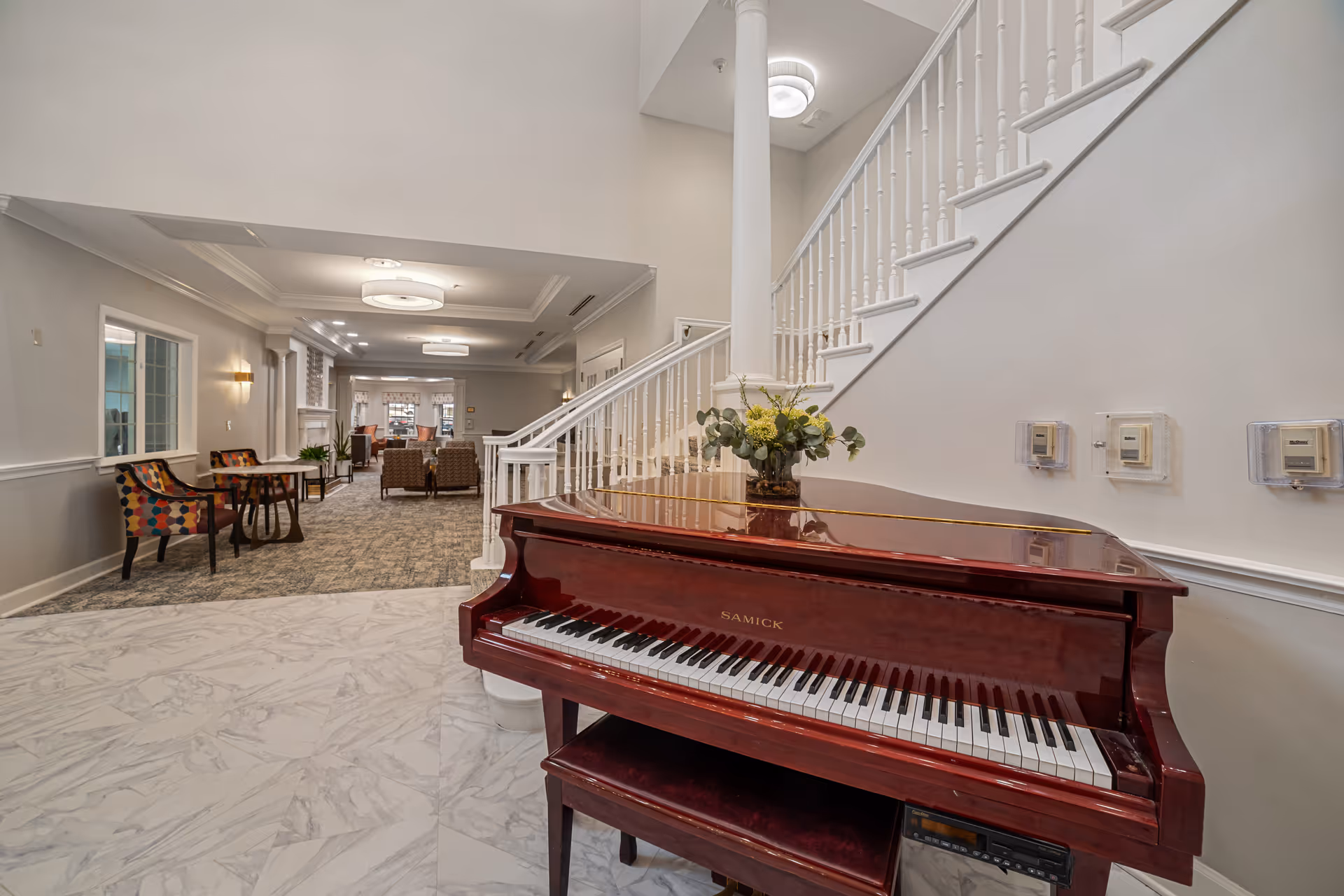 Interior view of a senior living facility showing a polished wooden grand piano with a flower arrangement on top, positioned near a white staircase with spindles. The room has light-colored walls, marble flooring near the piano, and carpeted flooring further inside. There are chairs and tables along the left wall and a seating area with armchairs in the background under ceiling lights.