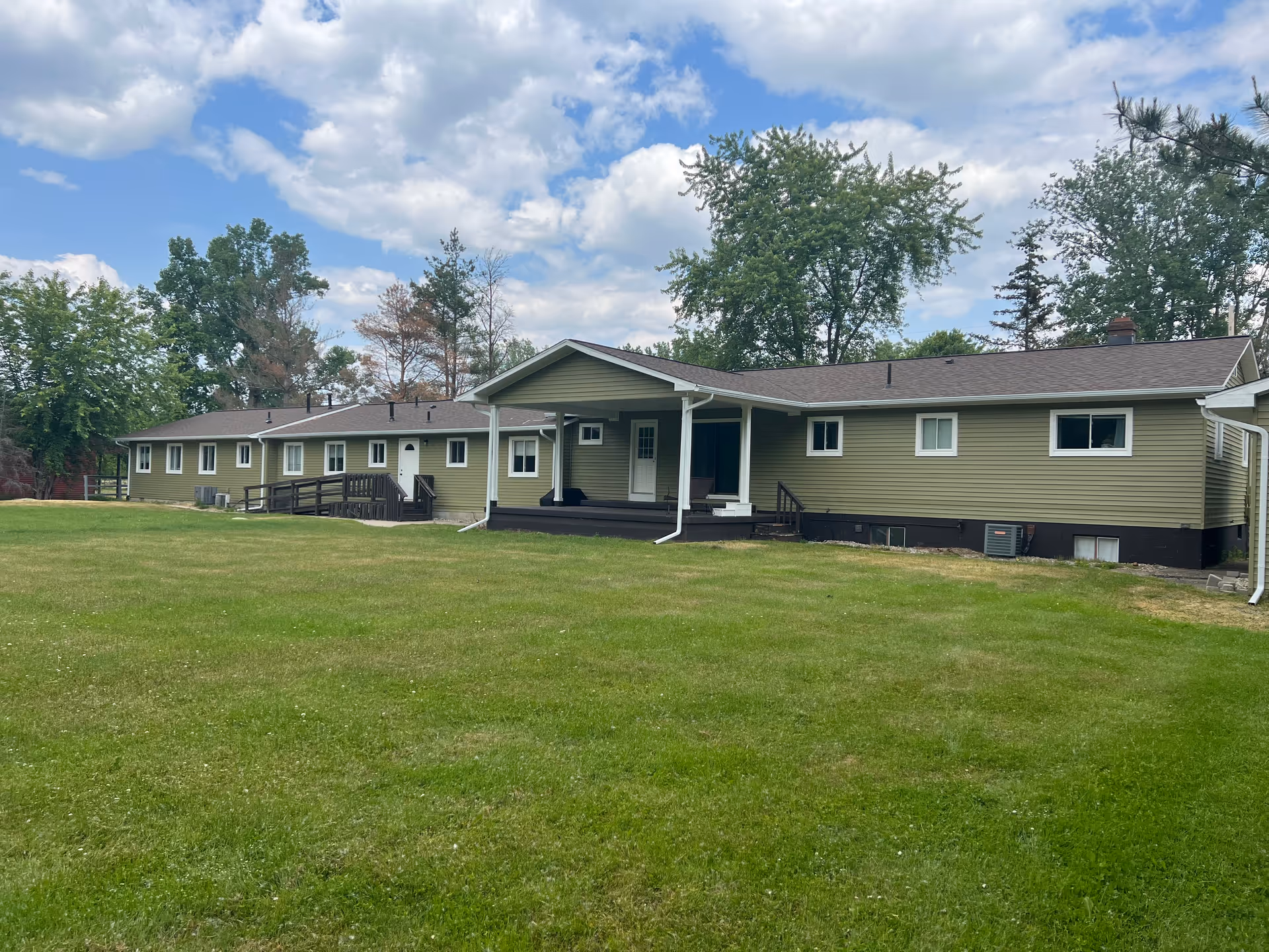 A single-story building with green siding and a brown roof, surrounded by a large grassy lawn and trees in the background under a partly cloudy sky.